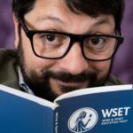Uomo con barba e occhiali legge un libro blu del WSET Wine & Spirit Education Trust