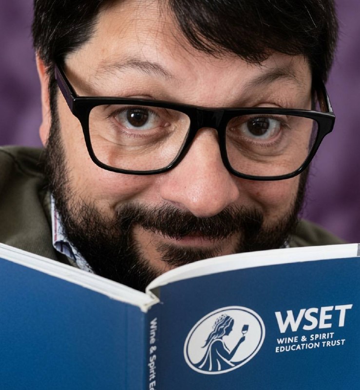 Uomo con barba e occhiali legge un libro blu del WSET Wine & Spirit Education Trust