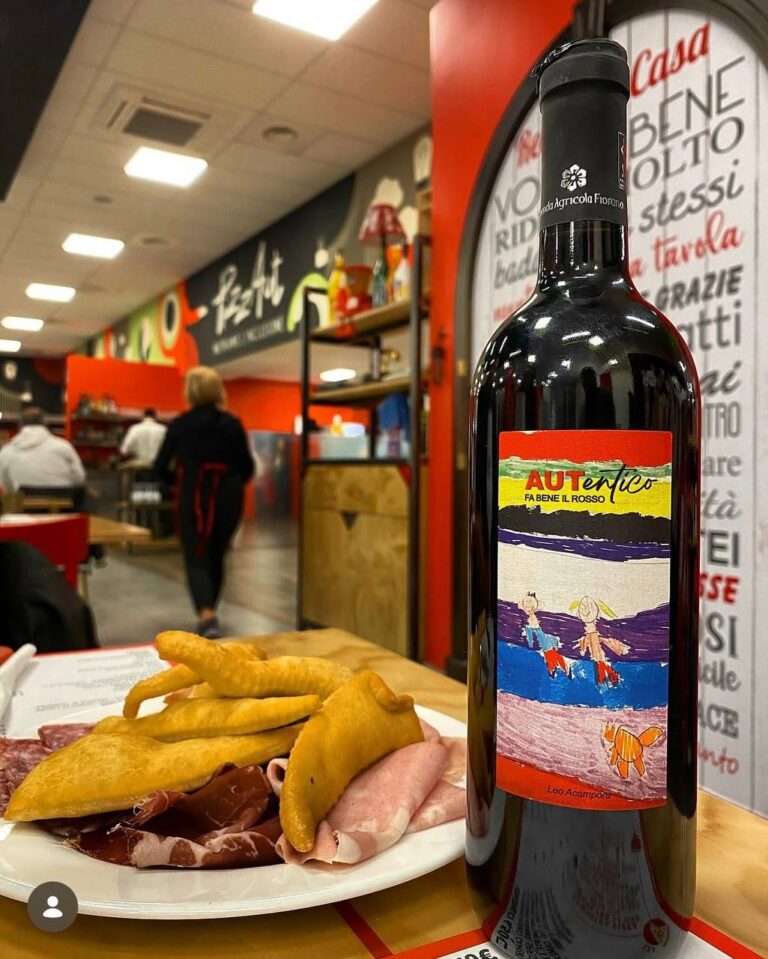 Bottiglia di vino Autentico con etichetta colorata accanto a un piatto di prosciutto e pane tostato in una pizzeria