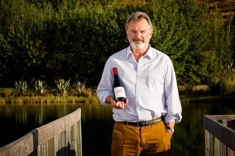 Sam Neill in piedi su una passerella di legno affacciata su un lago, con bottiglie di vino in mano, alberi verdi sullo sfondo