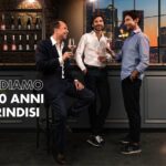 Tre uomini in abbigliamento business brindano con calici di vino rosato in una lounge con skyline cittadino sullo sfondo