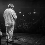 Cantautore di spalle sul palco davanti al pubblico, con una fiaschetta di vino accanto