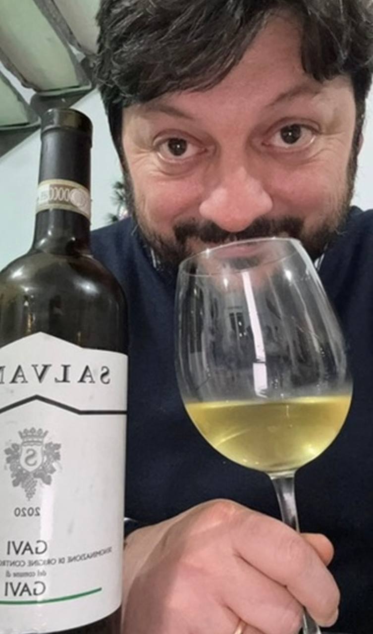 Uomo con barba assaggia vino bianco da un calice, con bottiglia di Savías sullo sfondo