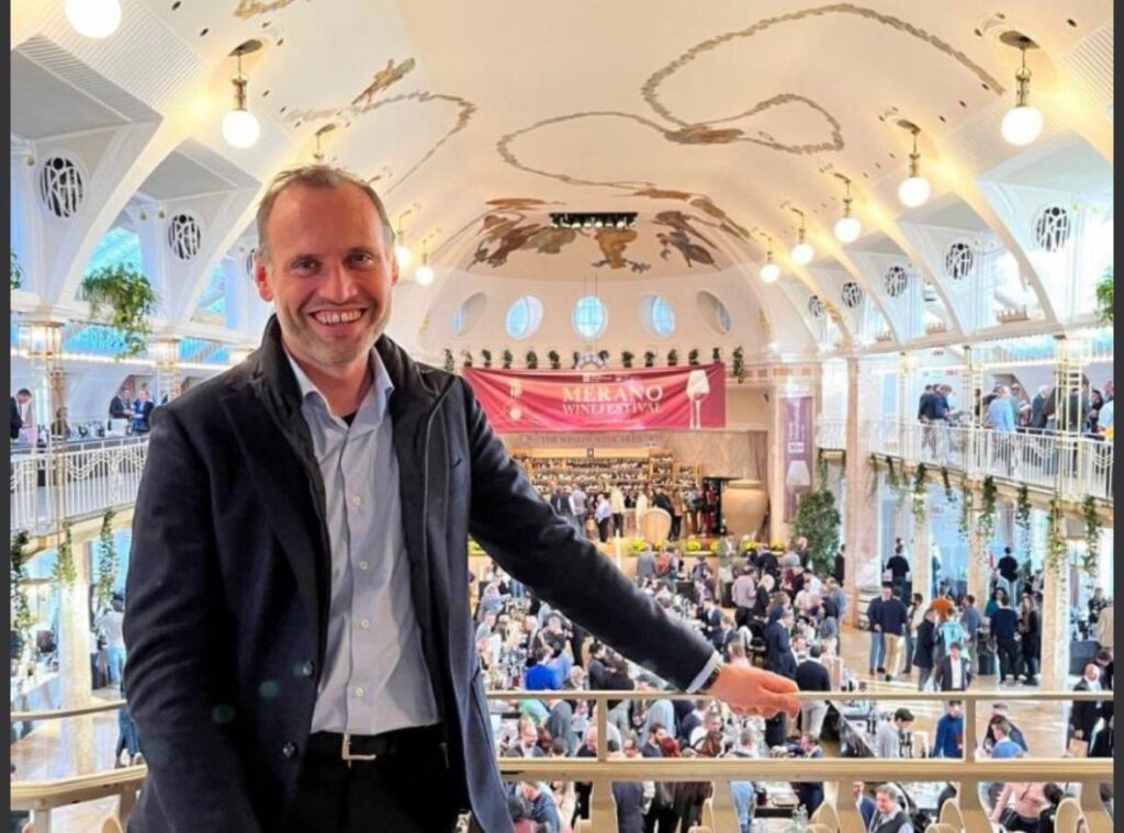 Uomo sorride alla telecamera in una grande sala con cupola, durante il Merano WineFestival con folle di visitatori sullo sfondo