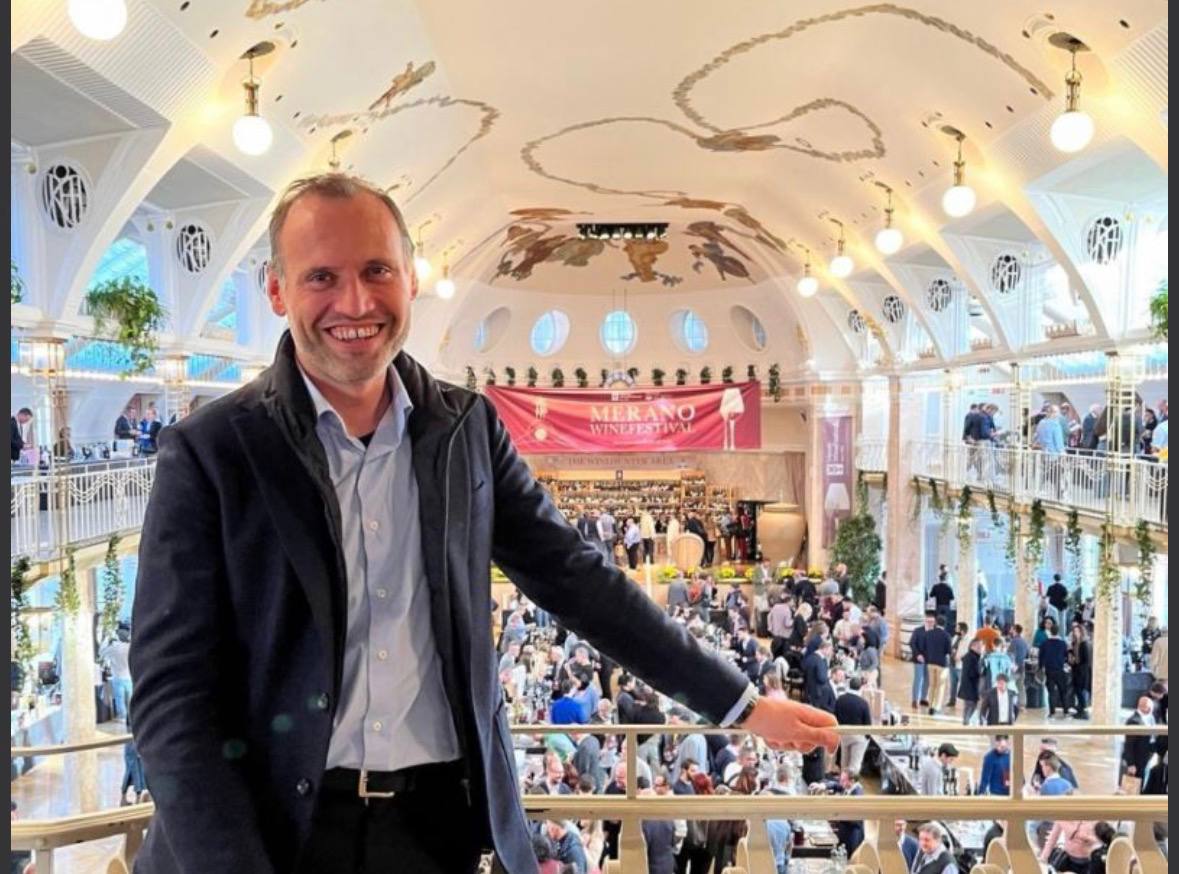 Uomo sorride alla telecamera in una grande sala con cupola, durante il Merano WineFestival con folle di visitatori sullo sfondo